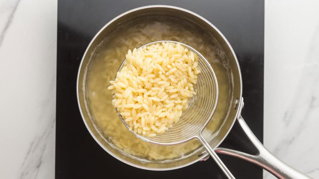 Cooked orzo being lifted from a pot of boiling water with a wire skimmer
