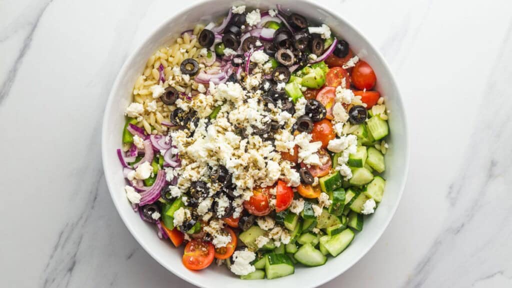 Greek orzo salad ingredients in a bowl with cooked orzo, cherry tomatoes, cucumber, red onion, olives, and crumbled feta
