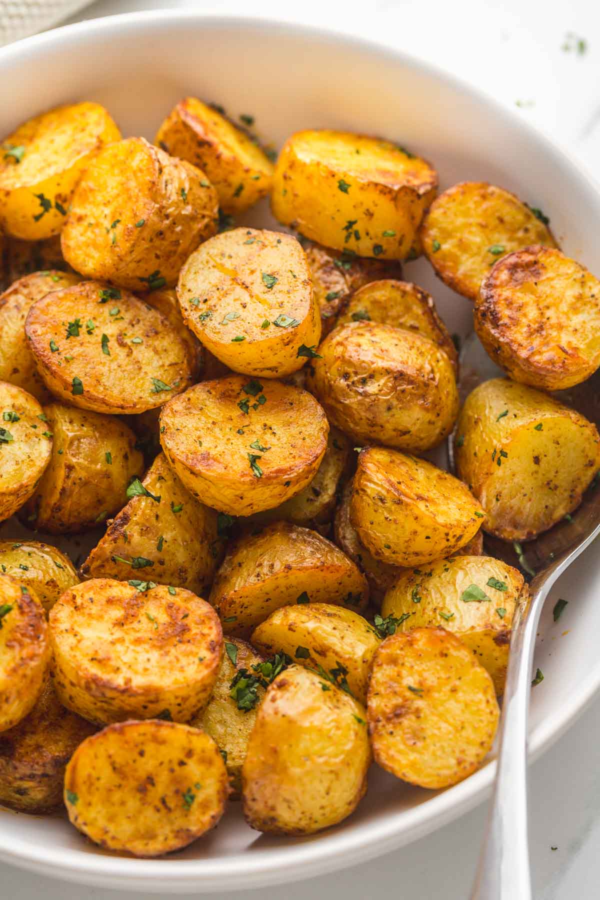 Close up shot of roasted potatoes in a white bowl, with a serving spoon on the side.