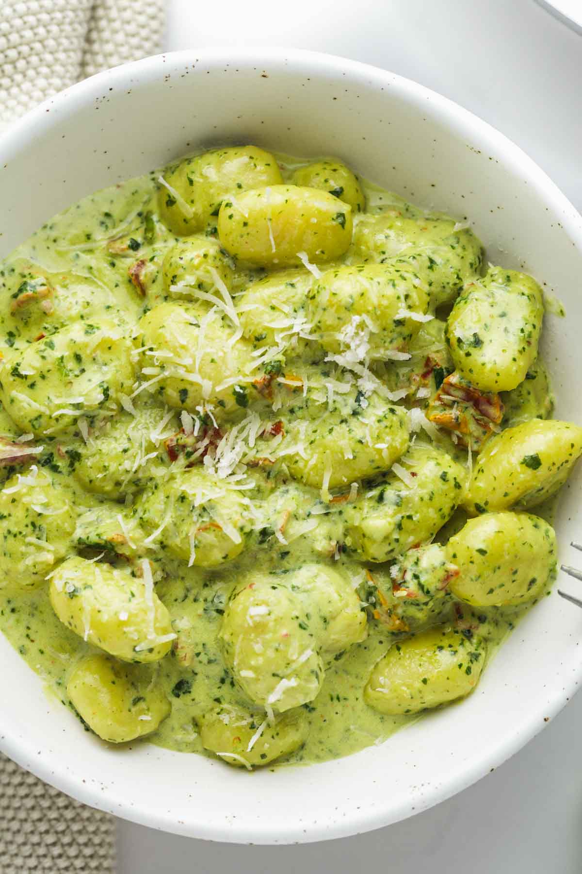 Creamy pesto gnocchi served in a white speckled bowl, with grated parmesan cheese on top.