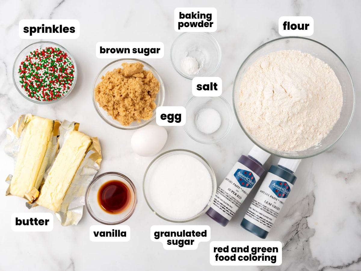 Overhead view of ingredients for Christmas pinwheel cookies, including butter, brown sugar, granulated sugar, vanilla, an egg, all-purpose flour, baking powder, salt, red and green gel food coloring, and festive sprinkles, all neatly arranged on a white marble surface.