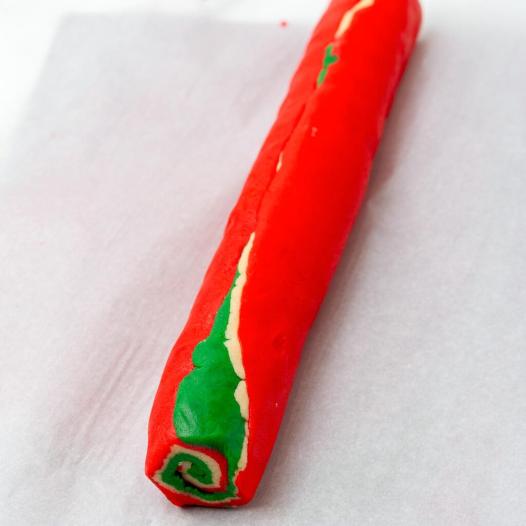 A rolled log of layered red, green, and white dough on parchment paper, showing a spiral pattern at the end, ready to be chilled for Christmas pinwheel cookies.