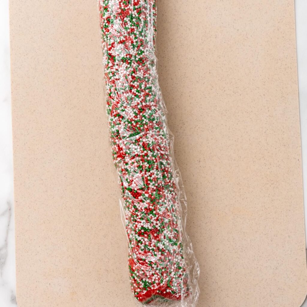 A log of layered red, green, and white dough coated in festive sprinkles, wrapped in plastic wrap, and ready to be chilled for Christmas pinwheel cookies.