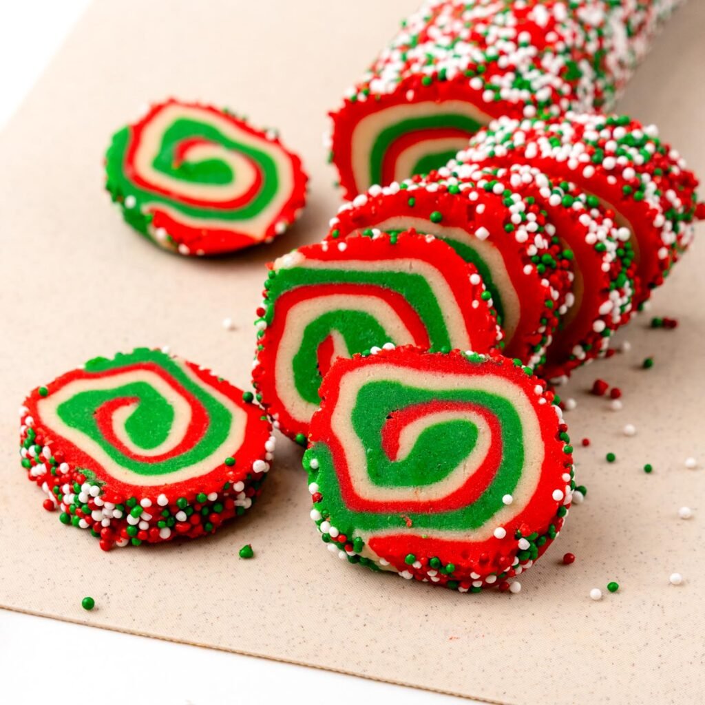 A log of Christmas pinwheel cookie dough coated in sprinkles, partially sliced into vibrant red, green, and white spiral cookies, ready for baking.