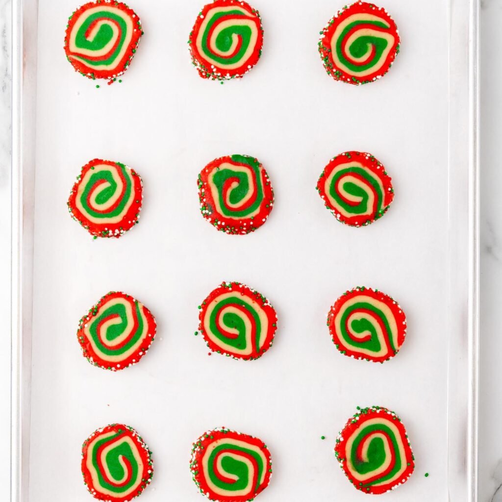 Sliced Christmas pinwheel cookies arranged on a parchment-lined baking sheet, showing vibrant red, green, and white spirals, ready to be baked.