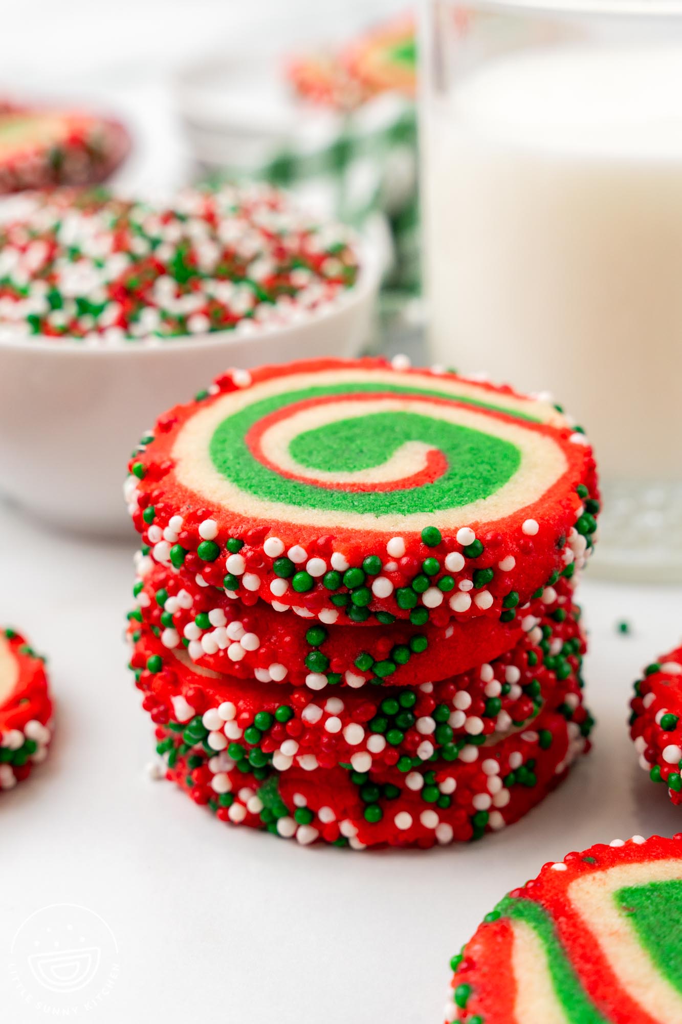 Stack of vibrant Christmas pinwheel cookies with red, green, and white swirls, coated in festive sprinkles, placed on a white surface with a glass of milk and bowls of sprinkles in the background.