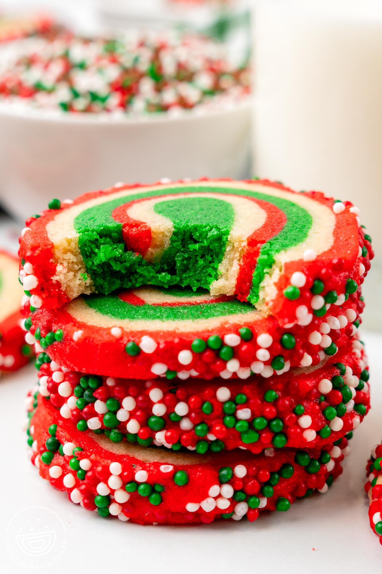 Stack of vibrant Christmas pinwheel cookies showing a bite shot with red, green, and white swirls, coated in festive sprinkles, placed on a white surface with a glass of milk and bowls of sprinkles in the background.