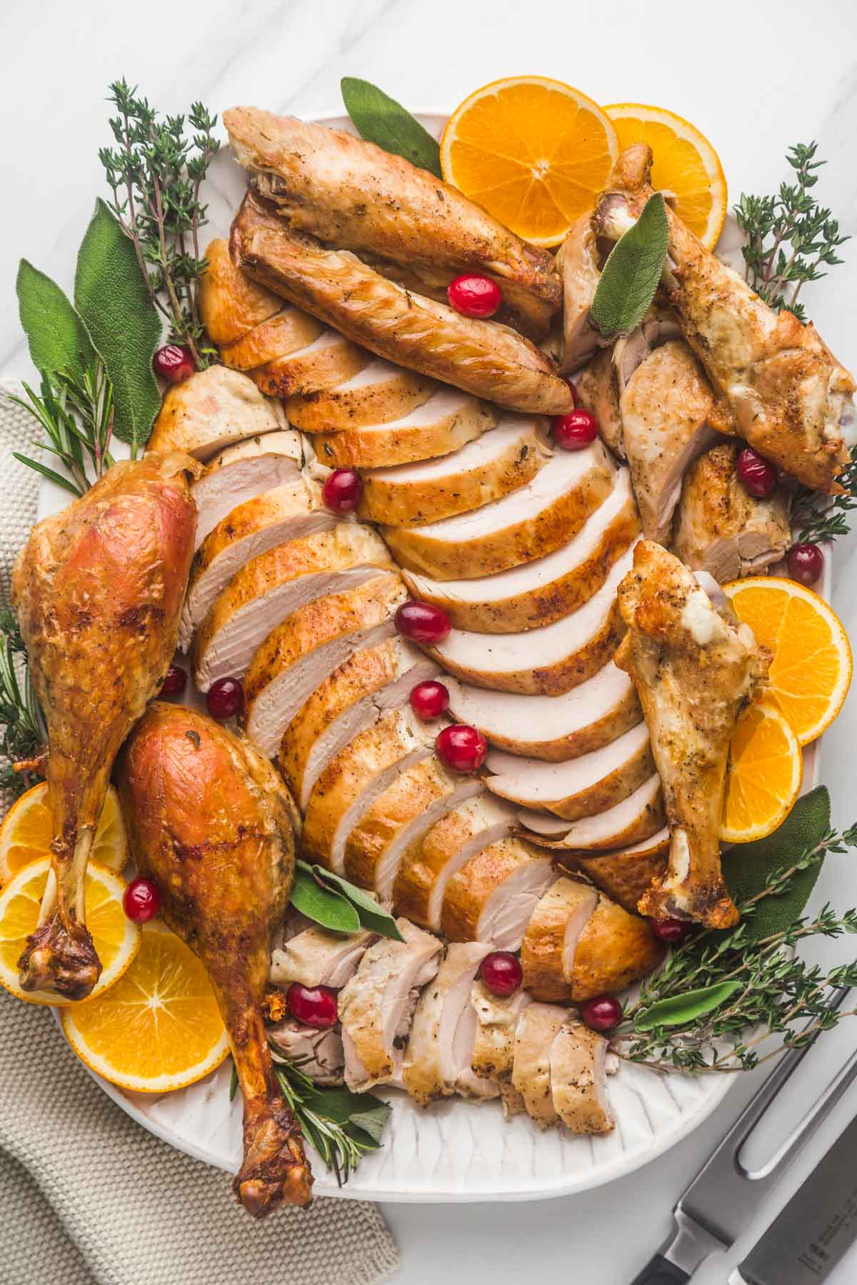 Carved and sliced turkey pieces served on a large white platter, with fresh herbs, orange slices, and fresh cranberries.
