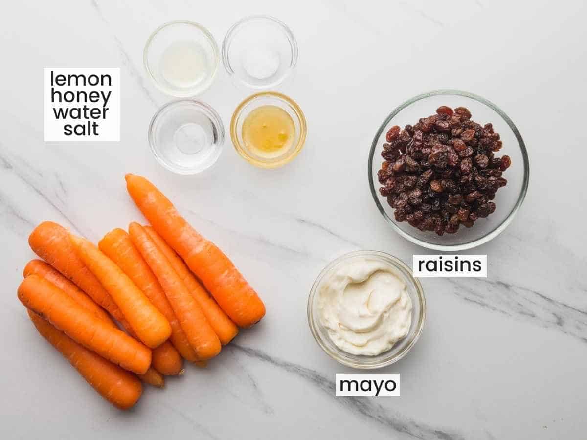 Overhead shot of ingredients needed to make a carrot salad including carrots, raisins, mayo, honey, lemon juice, water, and salt.