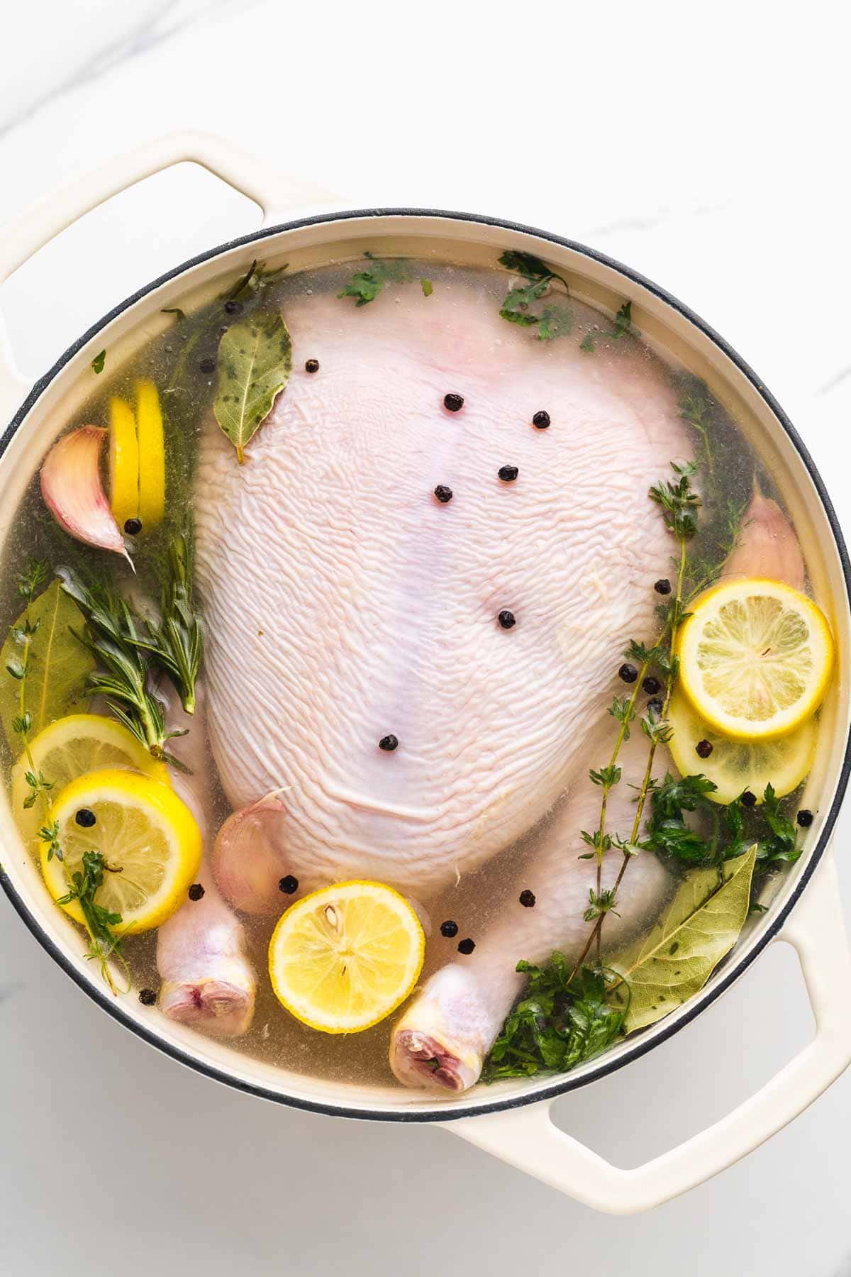 A whole chicken being brined in a large pot with slices of lemon, herbs, garlic cloves, and black peppercorns.