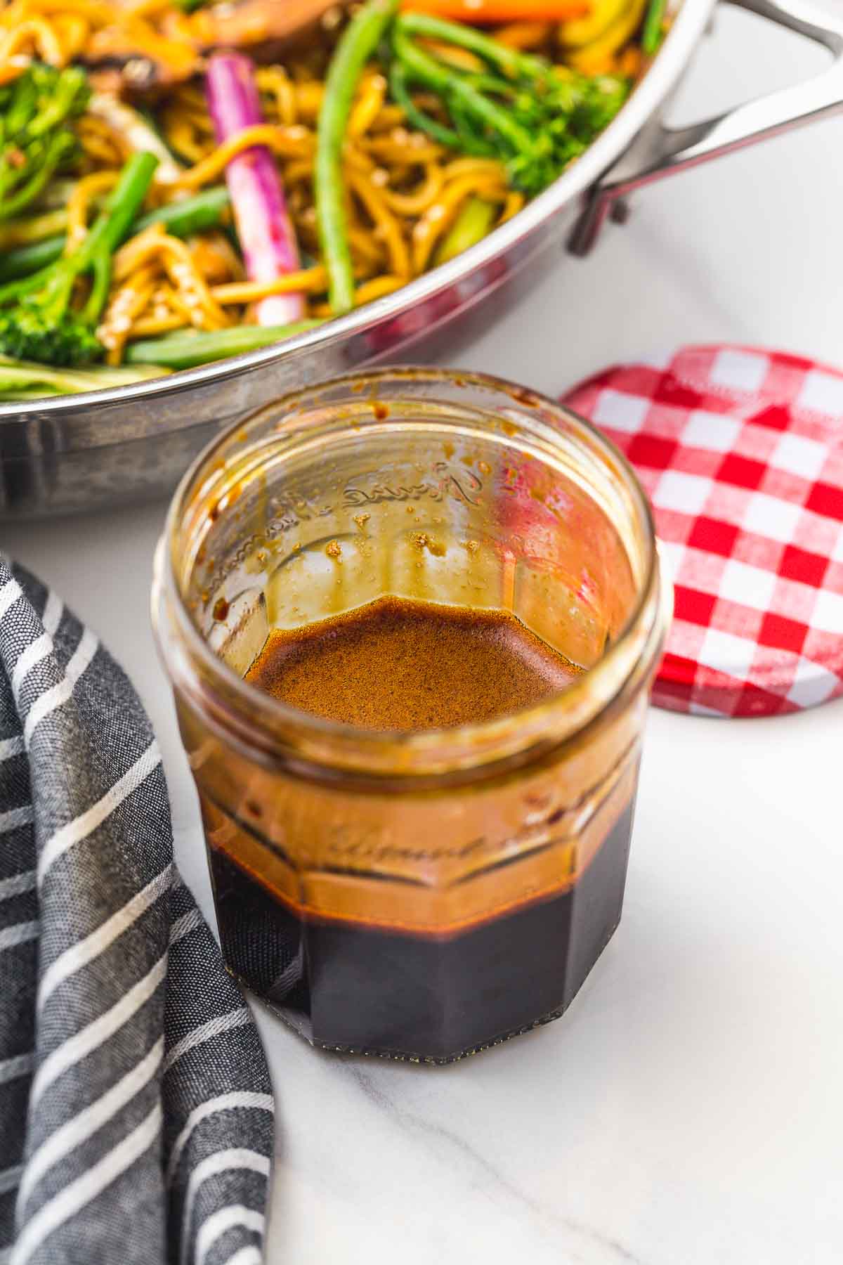 A glass jar with stir fry sauce, and a vegetable stir fry in the background.