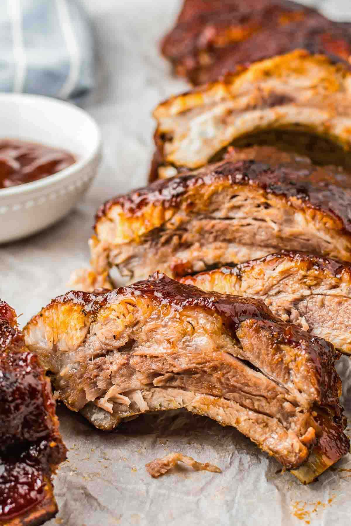 A close up of seperated barbecue baby back ribs served with additional BBQ sauce in a small bowl