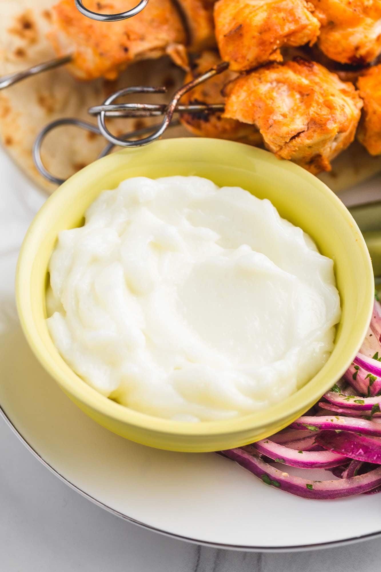 A small yellow bowl of garlic sauce, and shish tawook on skewers in the background.