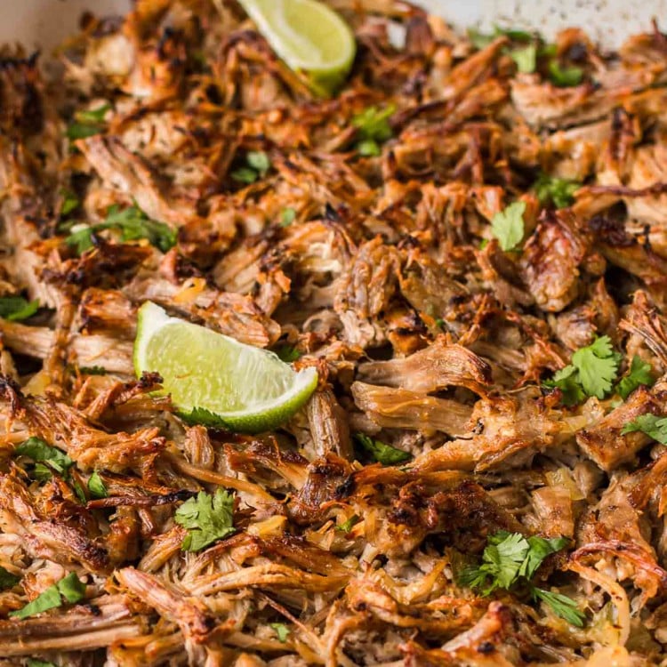 Crispy shredded carnitas in a pan with lime wedges and fresh cilantro