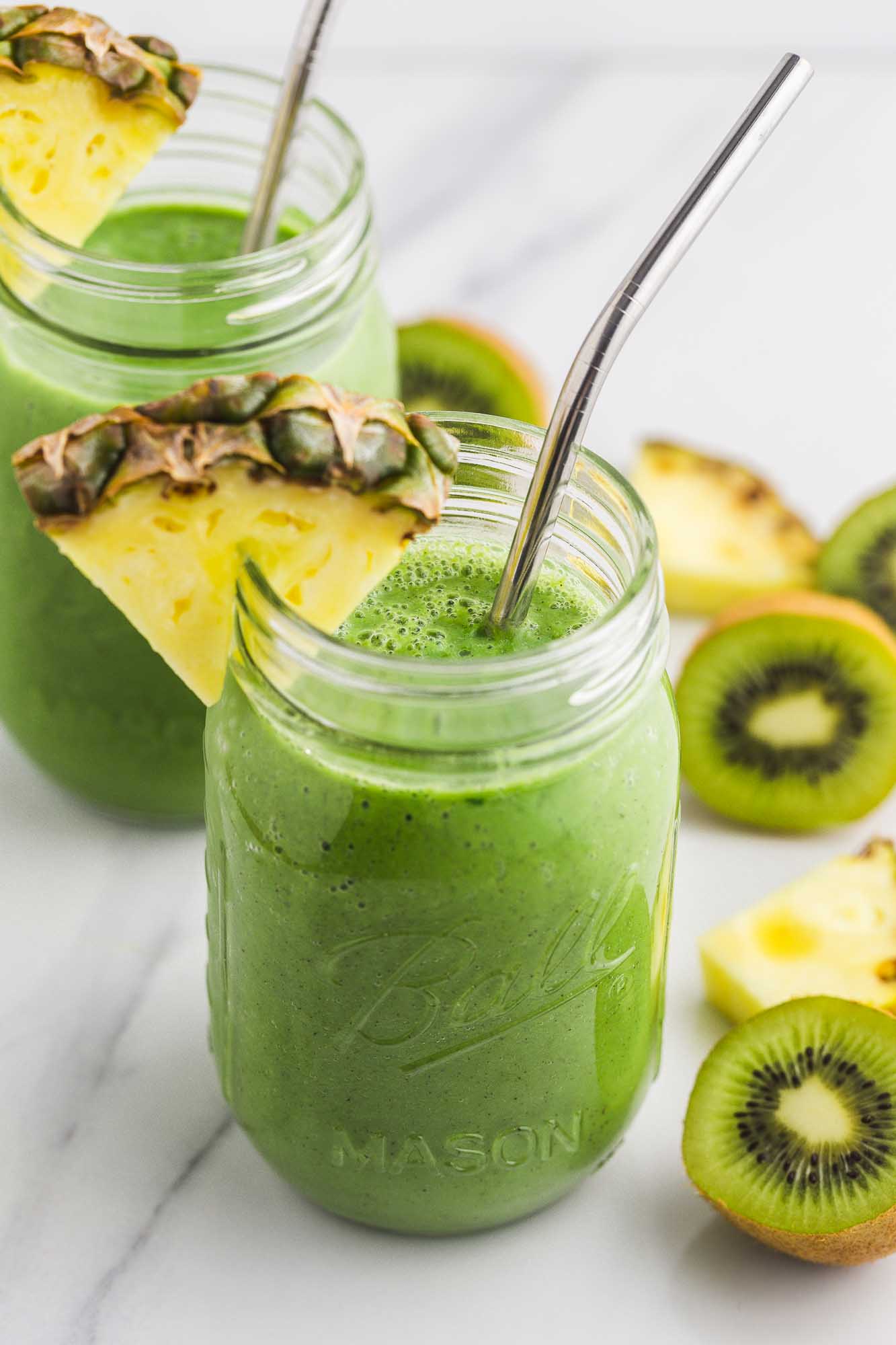 Spinach smoothie served in mason jars, with fresh pineapple slices, halved kiwi, and metal straws