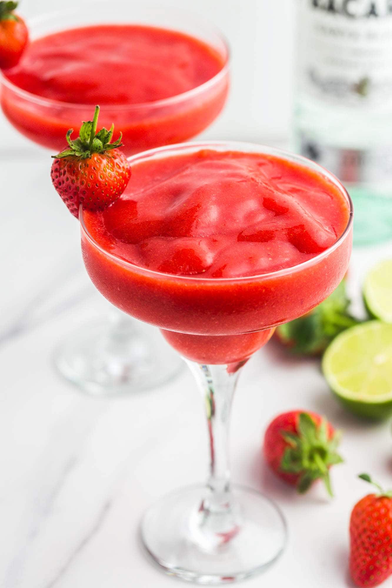 Strawberry Daiquiri in martini glasses, garnished with fresh strawberries. With a bottle of rum, fresh limes, and fresh strawberries in the background.