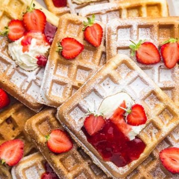 Strawberry waffles on a large white platter with a sprinkle of powdered sugar, topped with whipped cream, strawberry sauce, and fresh strawberries.
