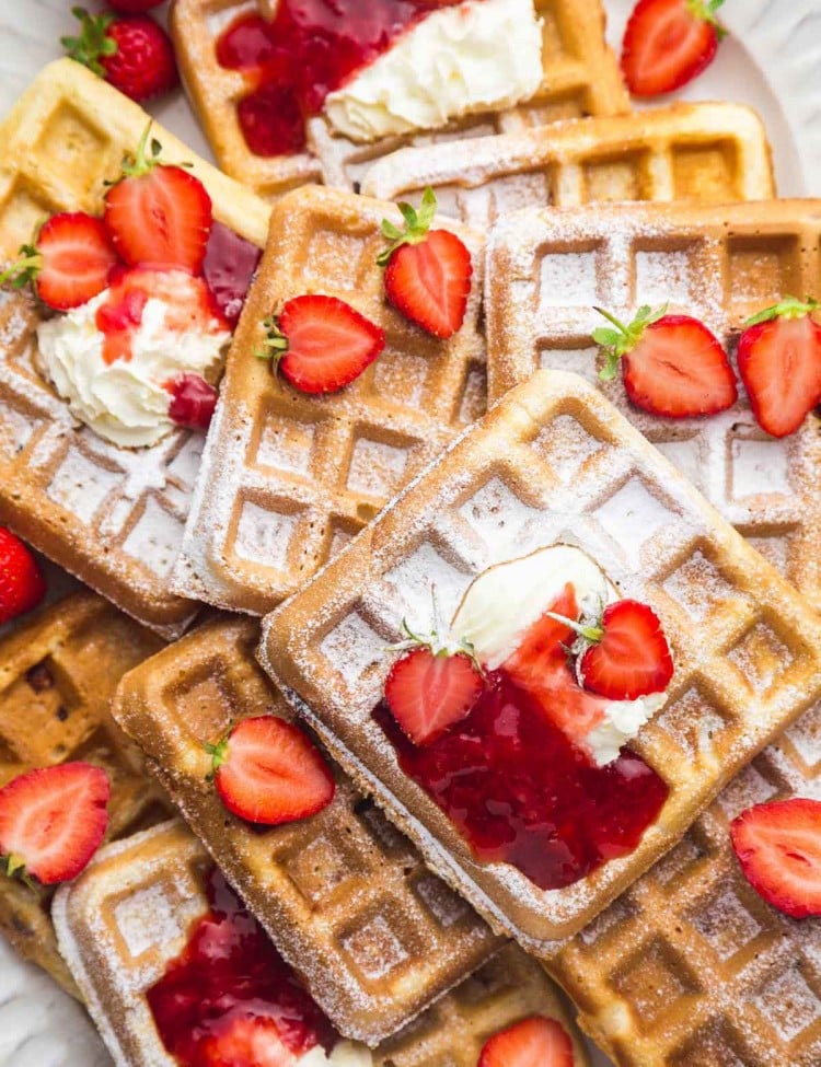 Strawberry waffles on a large white platter with a sprinkle of powdered sugar, topped with whipped cream, strawberry sauce, and fresh strawberries.