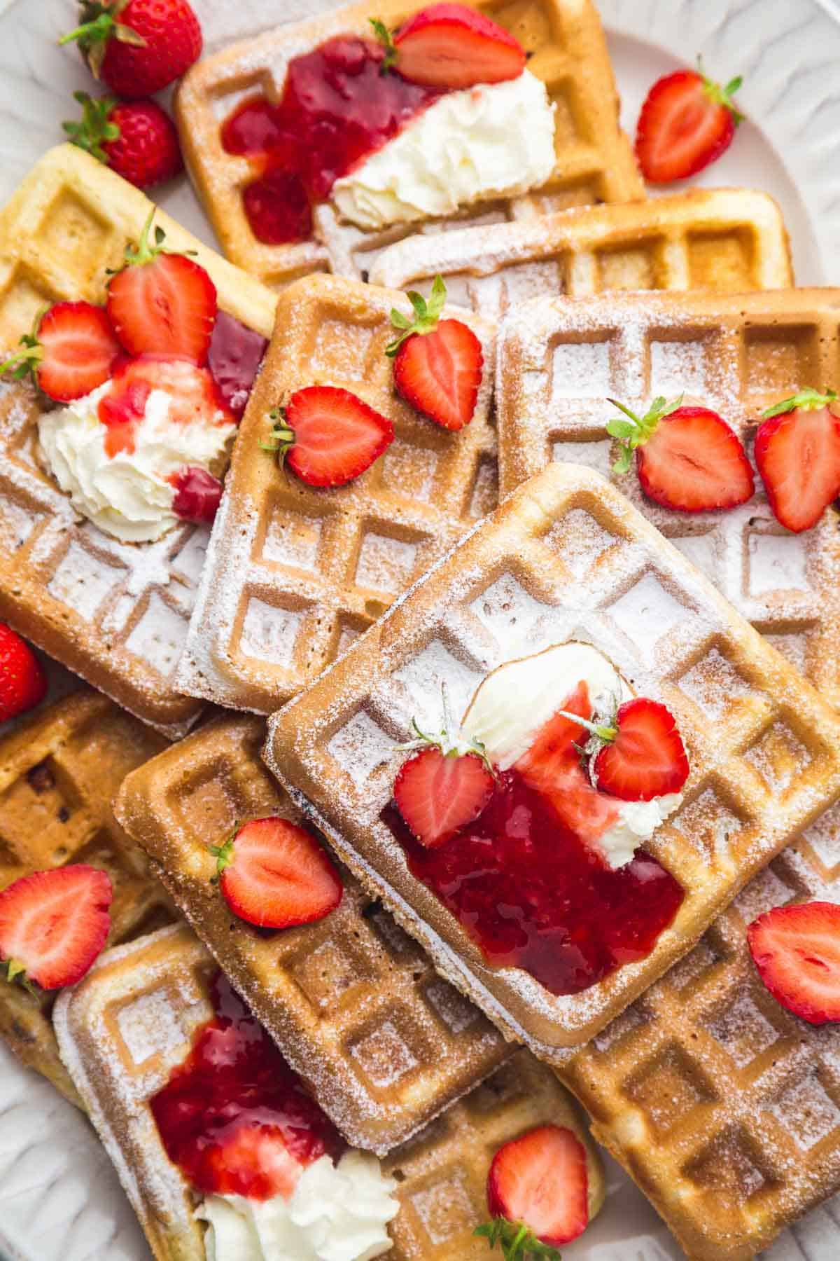 Strawberry waffles on a large white platter with a sprinkle of powdered sugar, topped with whipped cream, strawberry sauce, and fresh strawberries.