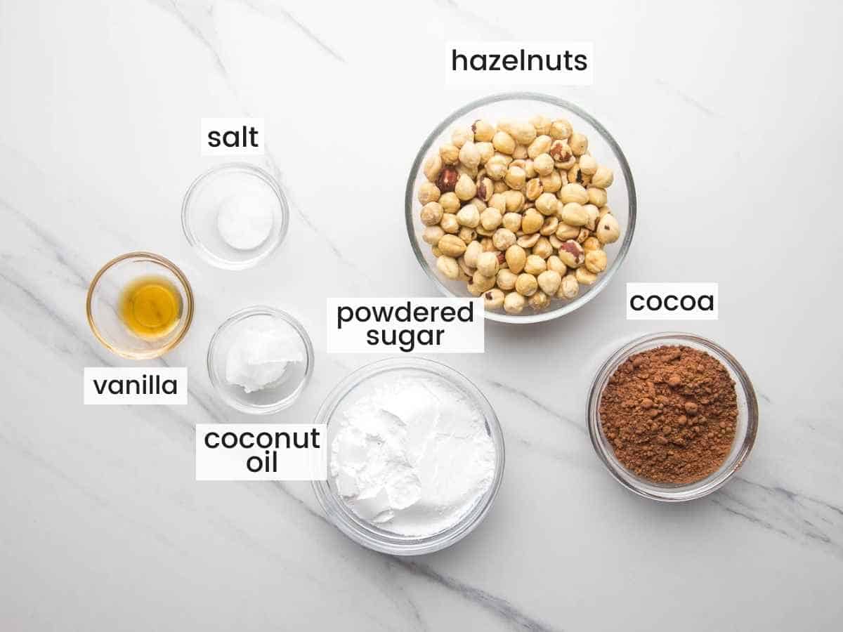 Ingredients for homemade nutella including hazelnuts, cocoa powder, powdered sugar, vanilla, coconut oil and salt.