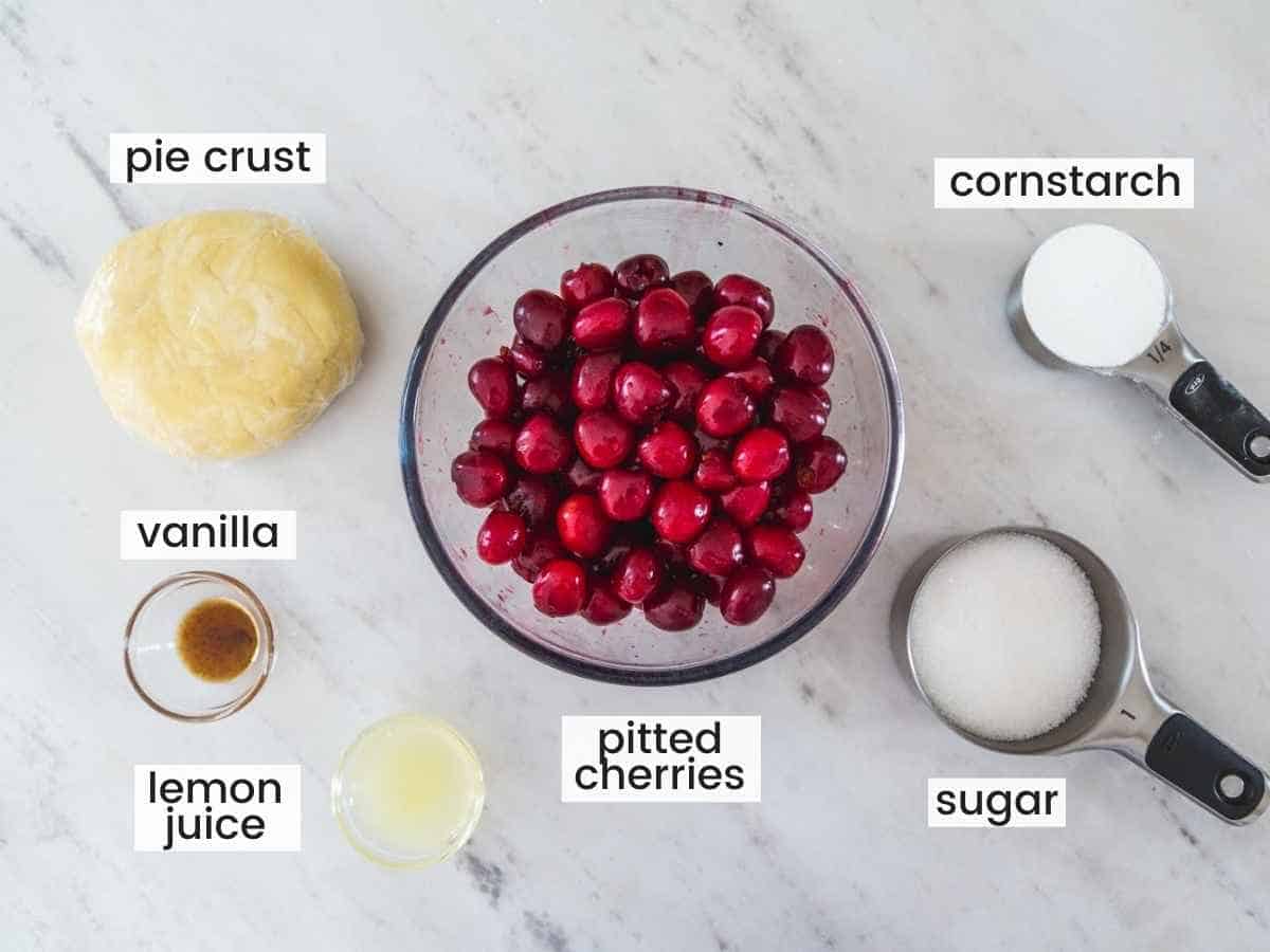 Ingredients needed to make a cherry pie including pitted cherries, pie crust dough, cornstarch, sugar, vanilla, and lemon juice.