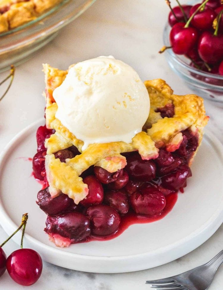 A slice of cherry pie served on a white dessert plate. Topped with vanilla ice cream.