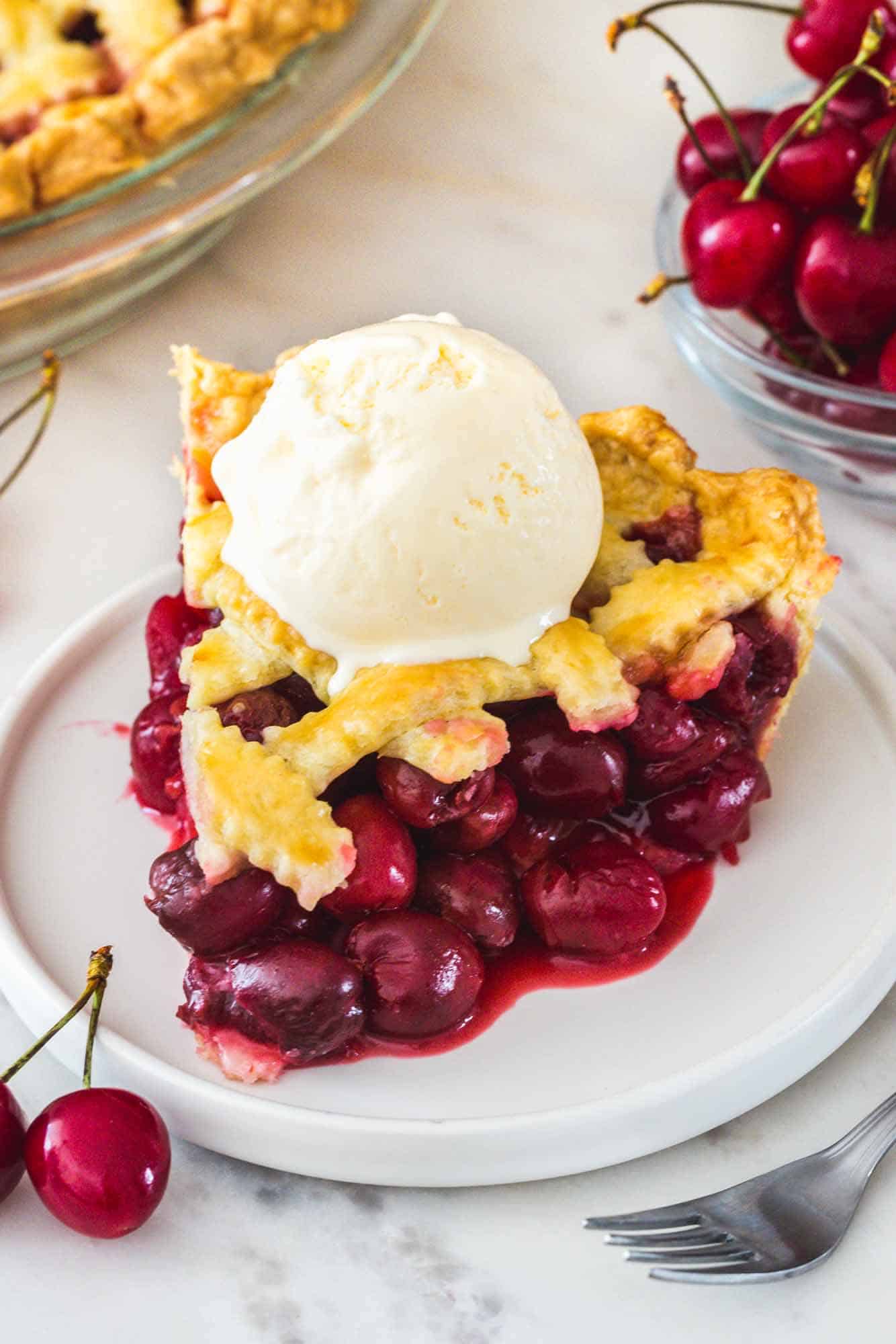 A slice of cherry pie served on a white dessert plate. Topped with vanilla ice cream.