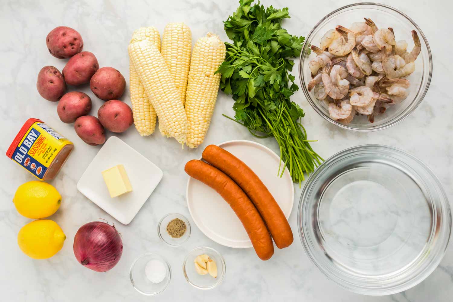 Ingredients needed to make shrimp boil including raw thawed shrimp, andouille sausage, red potatoes, lemons, old bay seasoning, red onion, corn, parsley and seasonings. 