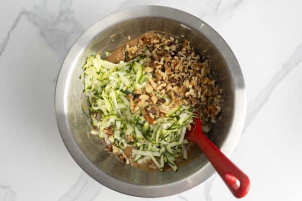 shredded zucchini and walnuts added to the cake mixture in a bowl, with a red spatula on the side.