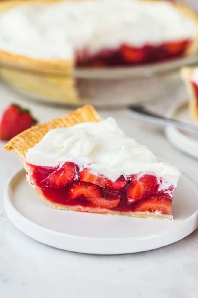 A slice of strawberry pie placed on a white plate, and a whole pie in the background