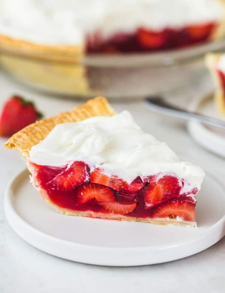 A slice of strawberry pie placed on a white plate, and a whole pie in the background