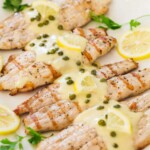 Grilled tilapia served on a platter, and topped with piccata sauce, capers and fresh lemon slices.