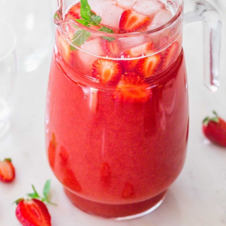 A glass pitcher with strawberry tea, ice cubes, fresh strawberries slices, and mint leaves garnish