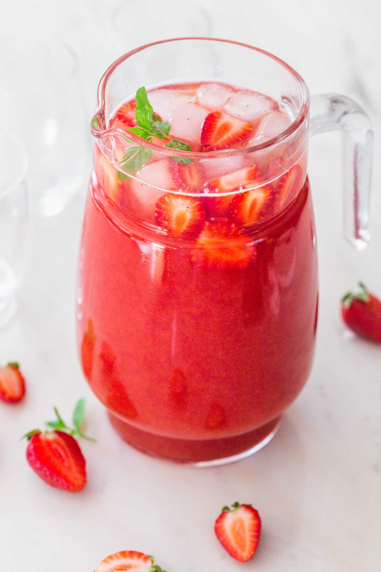 A glass pitcher with strawberry tea, ice cubes, fresh strawberries slices, and mint leaves garnish