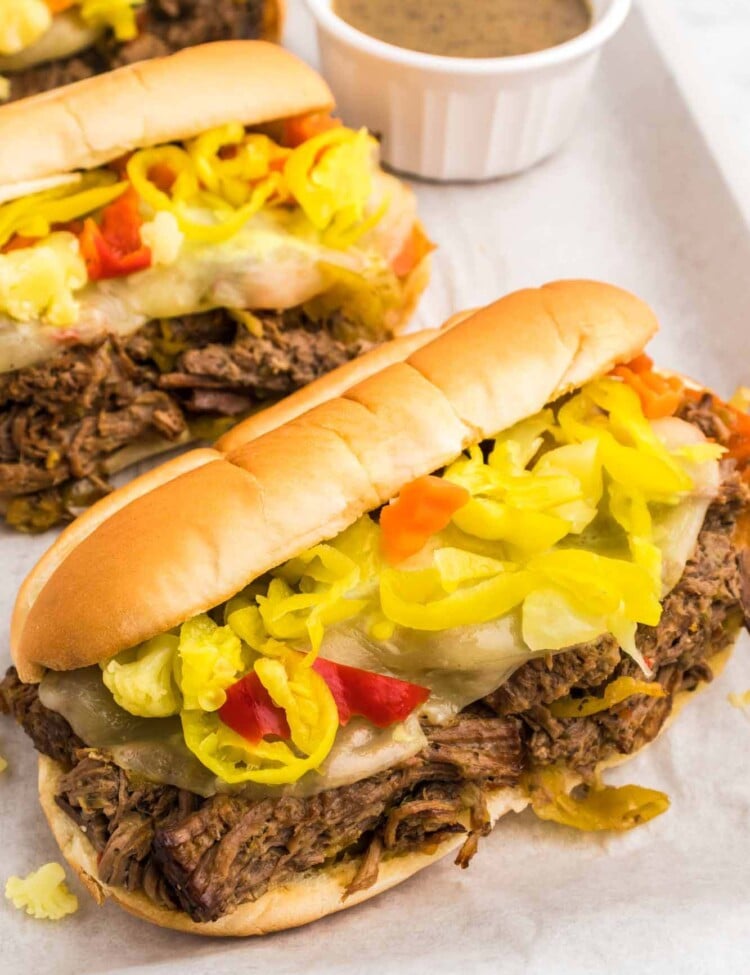 3 Italian beef sandwiches placed on parchment paper, and a little jus in a bowl for dipping.