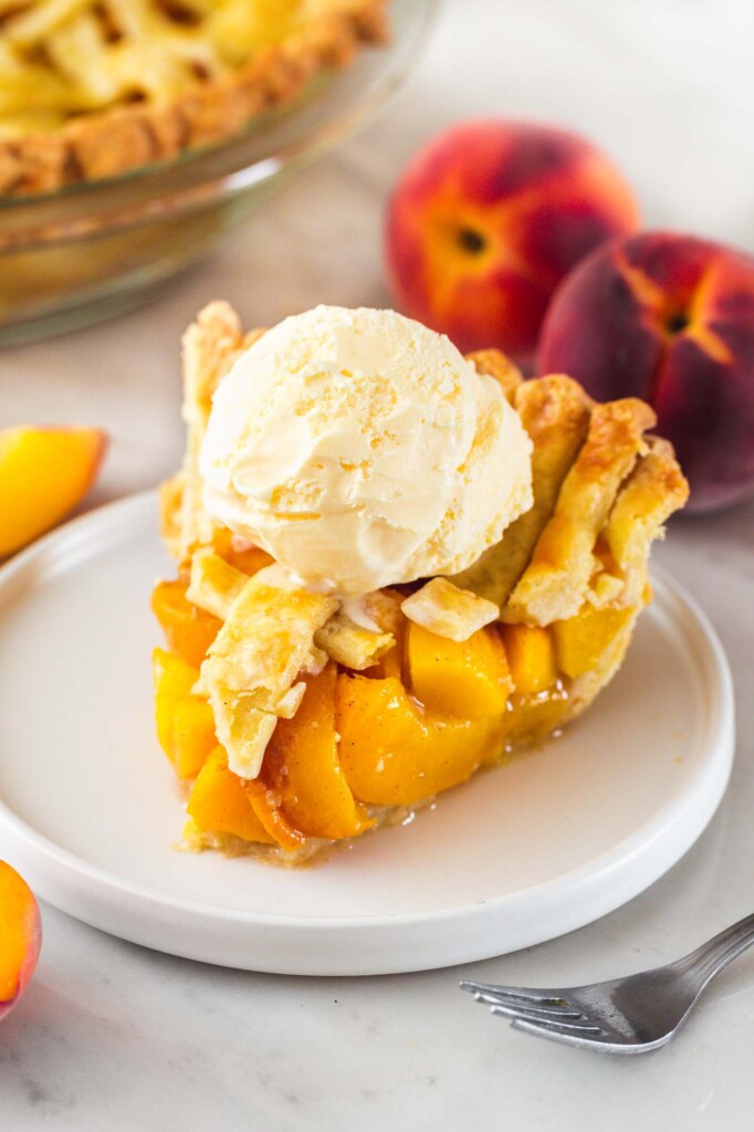 Slice of peach pie on a small white plate with a scoop of ice cream