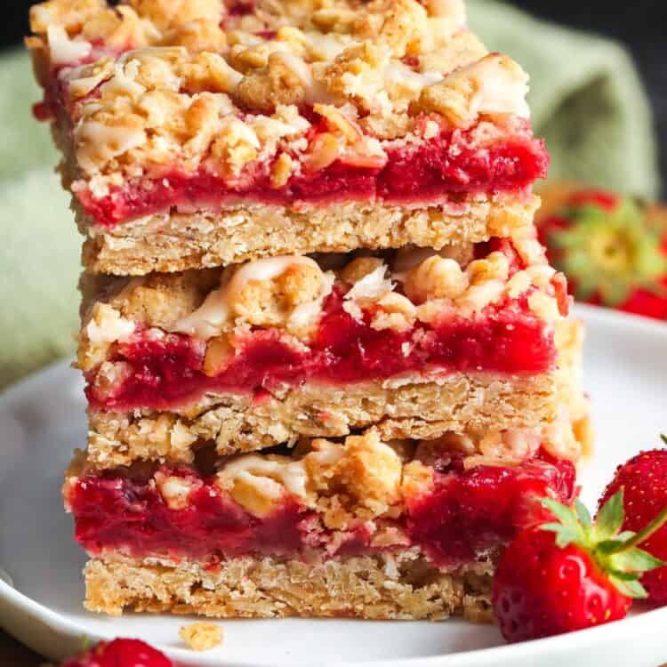 Strawberry oatmeal bars stacked on a white plate, topped with golden oat crumble and drizzled with vanilla glaze, with fresh strawberries on the side.