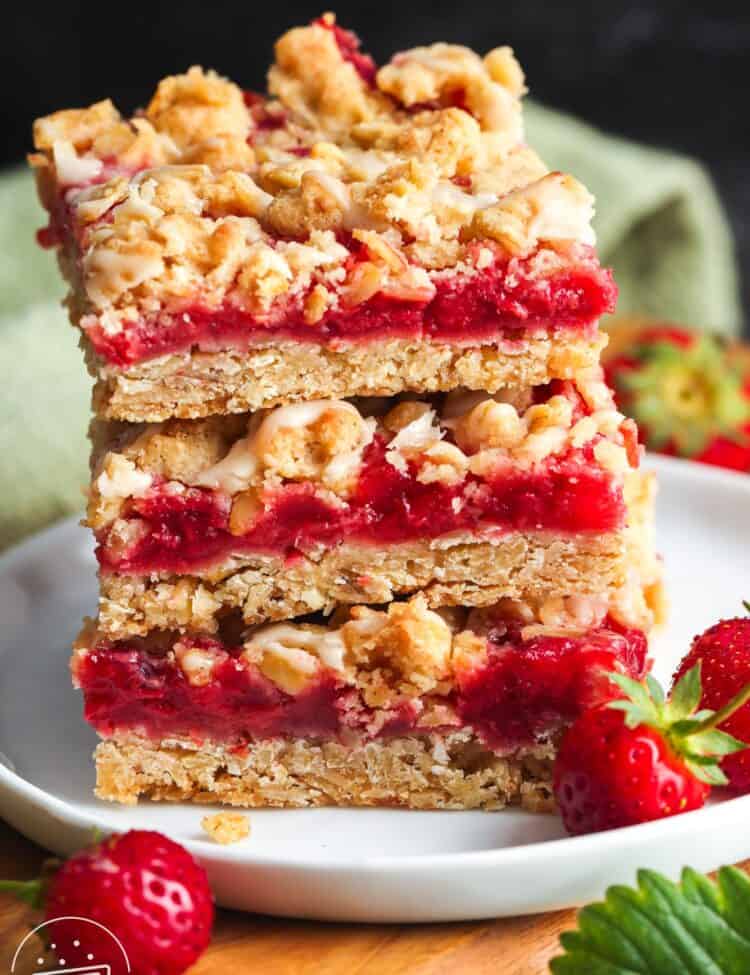 Strawberry oatmeal bars stacked on a white plate, topped with golden oat crumble and drizzled with vanilla glaze, with fresh strawberries on the side.