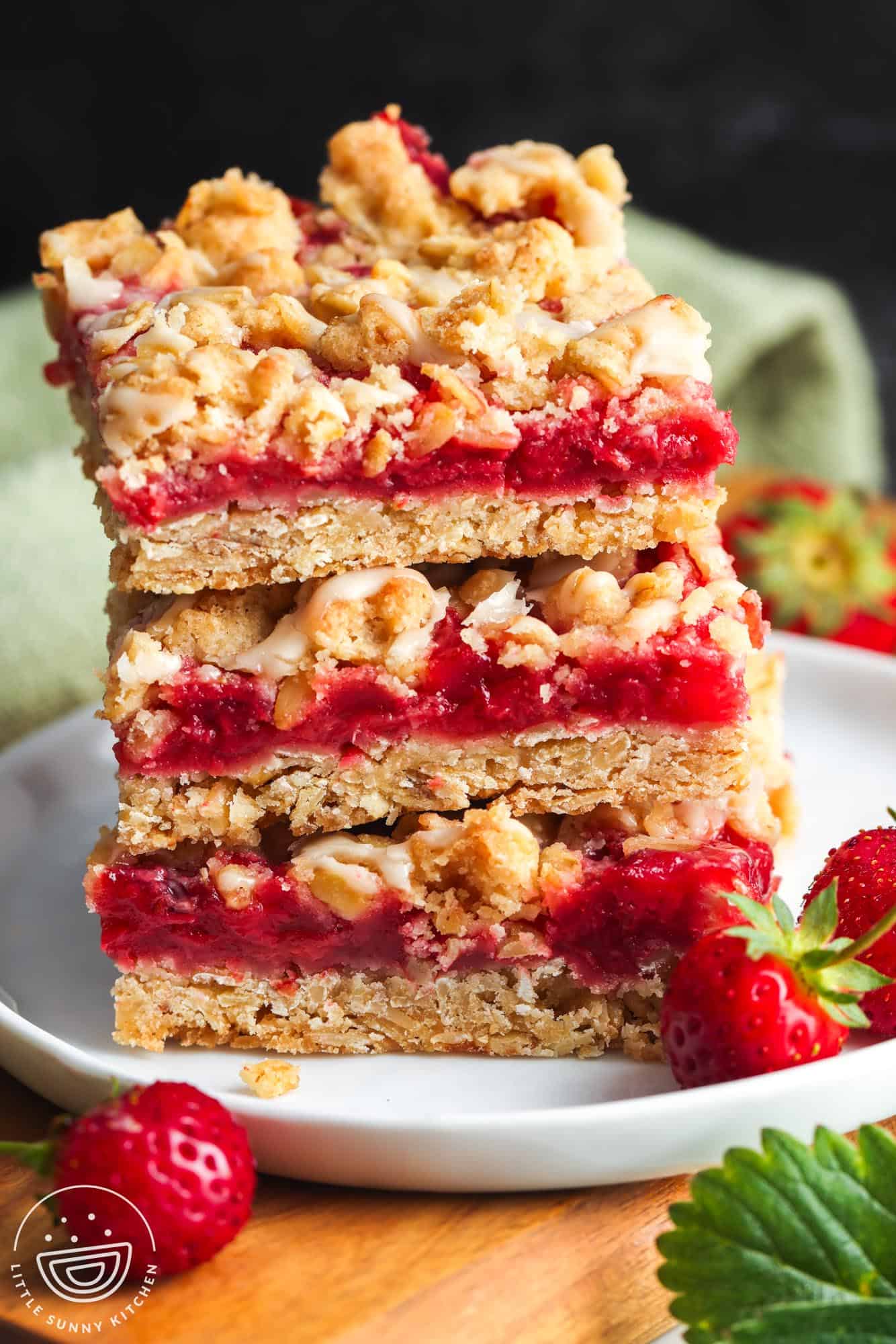 Strawberry oatmeal bars stacked on a white plate, topped with golden oat crumble and drizzled with vanilla glaze, with fresh strawberries on the side.