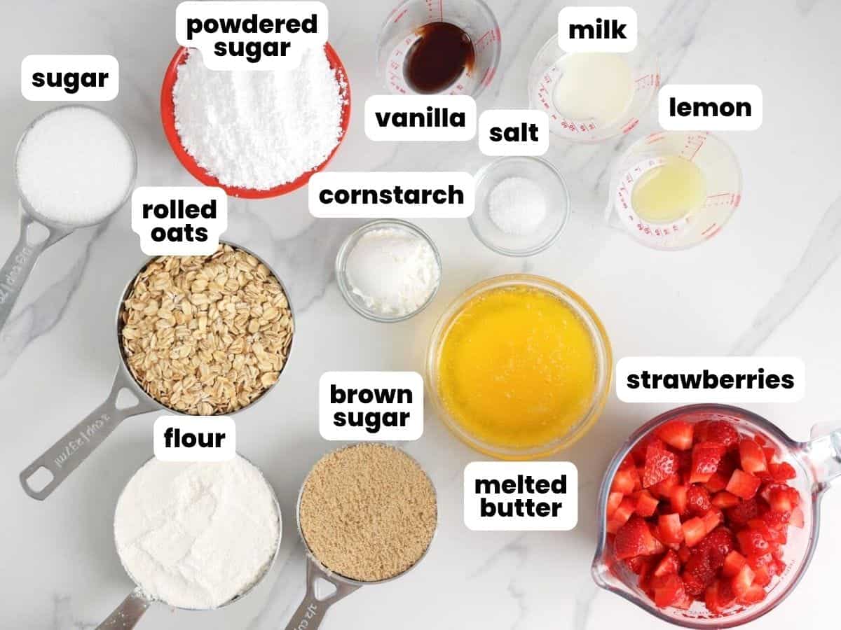 Overhead view of measured ingredients for strawberry oatmeal bars on a white marble surface, including bowls and measuring cups with fresh chopped strawberries, rolled oats, flour, brown sugar, granulated sugar, powdered sugar, melted butter, cornstarch, salt, lemon juice, milk, and vanilla extract, each labeled with text.