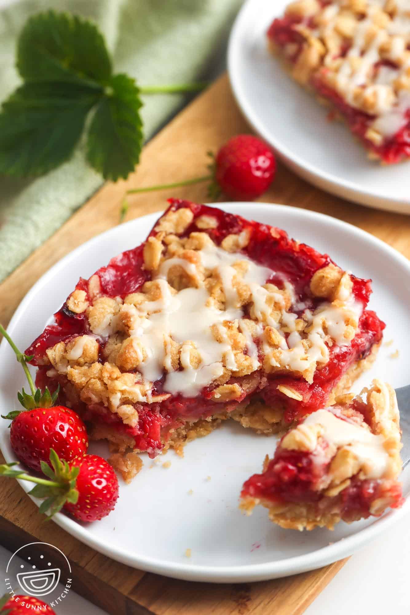 Close up of a strawberry oatmeal bar on a white plate, topped with golden oat crumble and drizzled with vanilla glaze, with fresh strawberries on the side and a bite taken out to show the jammy strawberry filling.