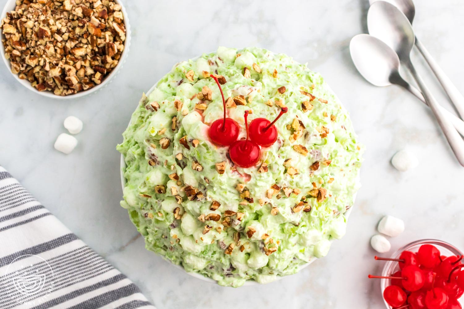 Horizontal image of an overhead shot of a pistachio dessert salad in a bowl, topped with maraschino cherries and pecan pieces. And a bowl of pecans on the side, and another bowl with maraschino cherries.