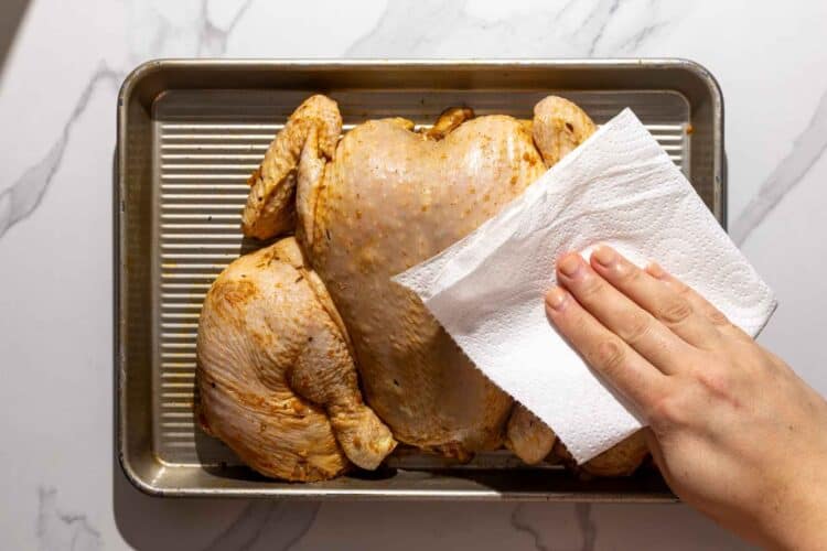 A hand pats a marinated spatchcocked chicken dry with a paper towel on a sheet pan.