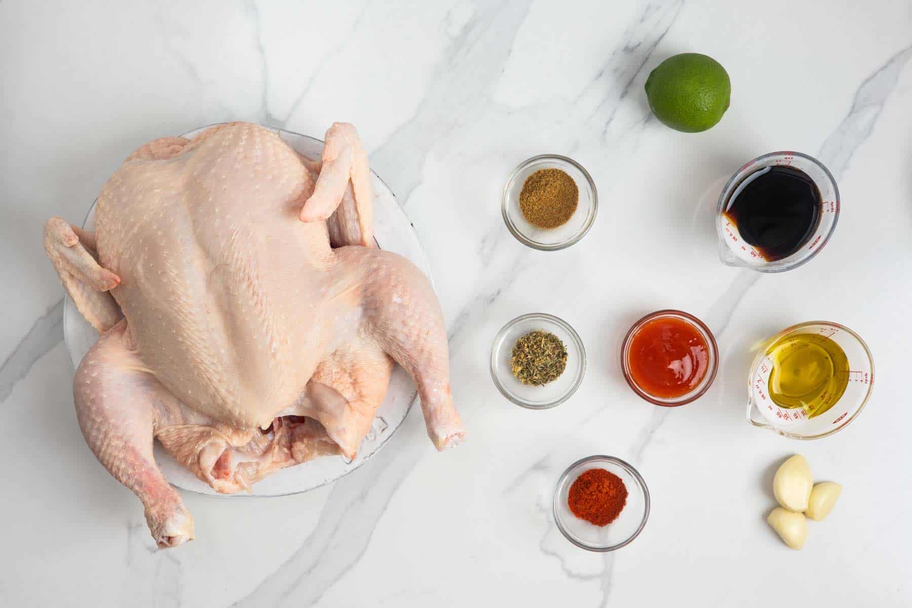 Ingredients for Peruvian chicken arranged with a whole chicken, spices, garlic, lime, oil, hot sauce, and soy sauce.