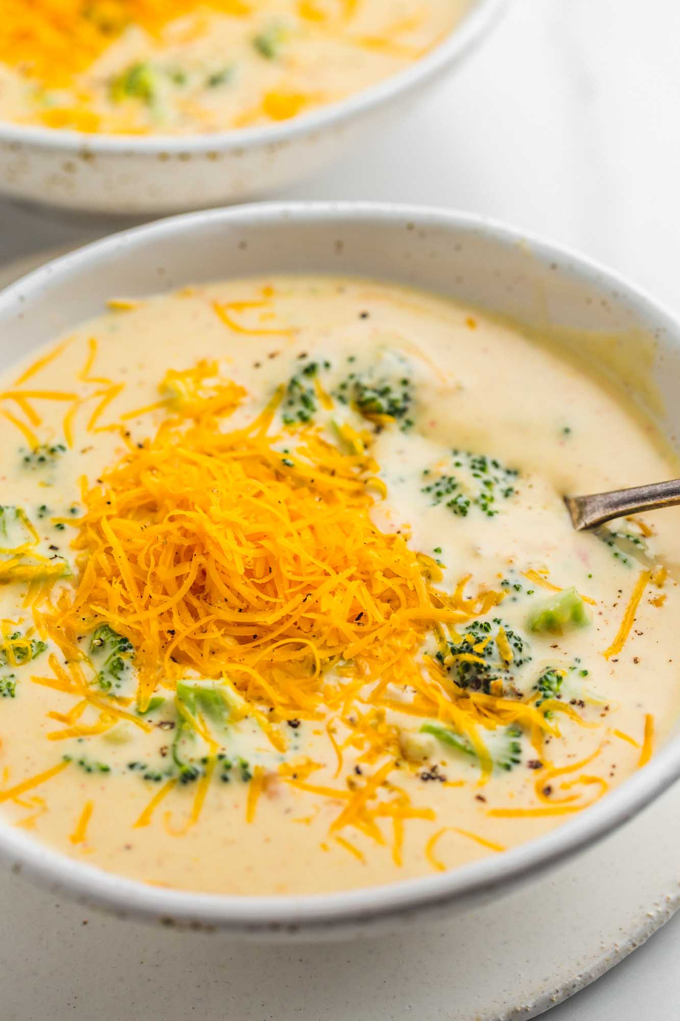 Creamy broccoli cheddar soup served in 2 white bowls with extra sharp cheddar cheese that is grated and added as a topping on the soup