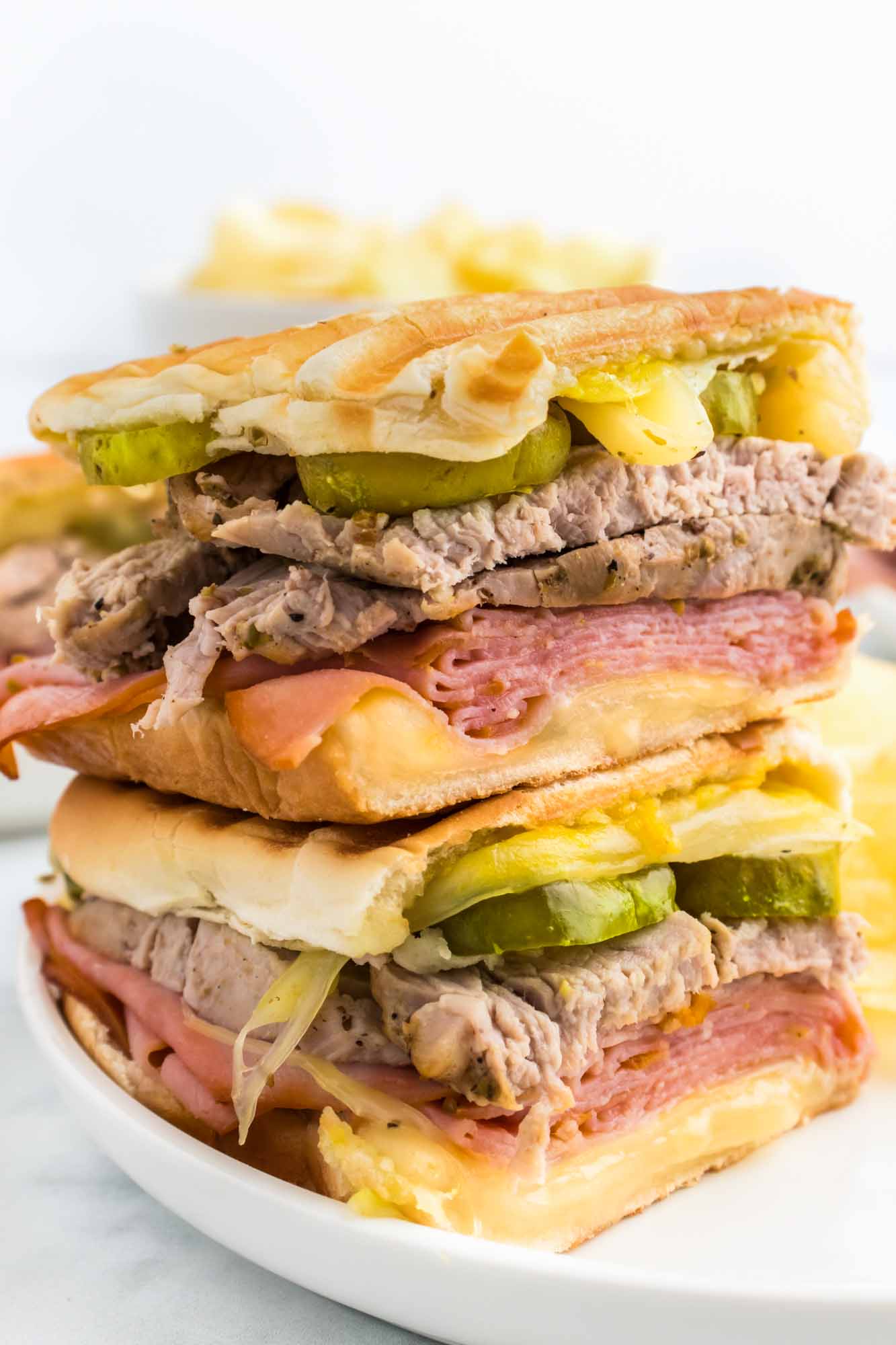 2 cuban sandwiches stacked, and served on a white plate with potato chips