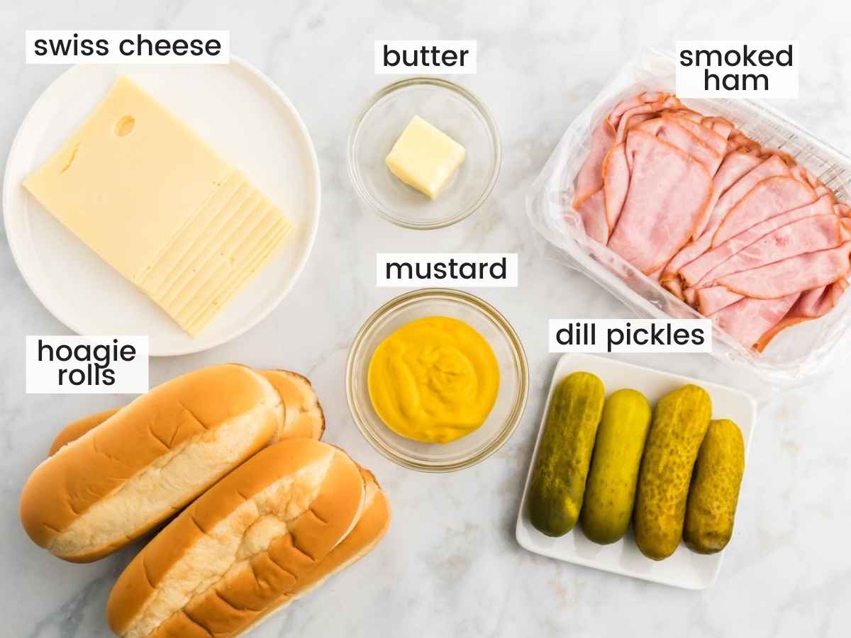 Ingredients needed to make a cuban sandwich including rolls, cheese, ham, pickles, mustard, and butter.