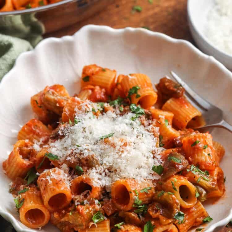 A white bowl filled with eggplant pasta topped with grated cheese and fresh parsley, with a fork on the side.