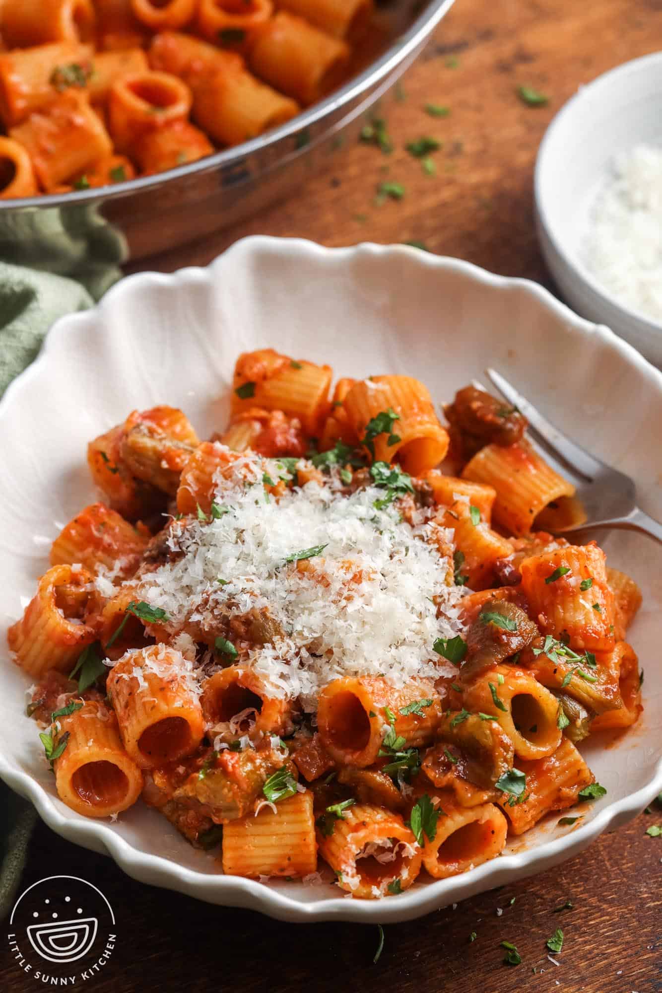A white bowl filled with eggplant pasta topped with grated cheese and fresh parsley, with a fork on the side.