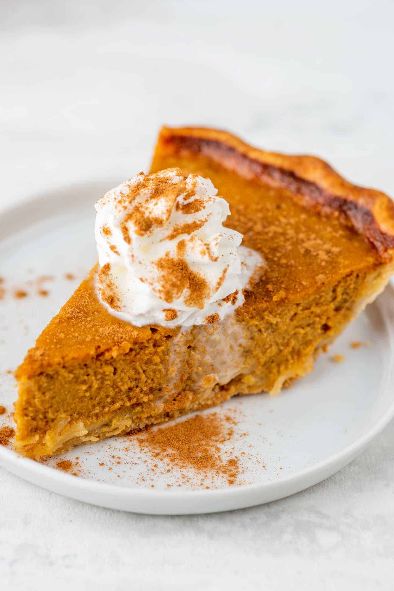 Slice of pumpkin pie on a white plate, topped with whipped cream and a sprinkling of ground cinnamon.